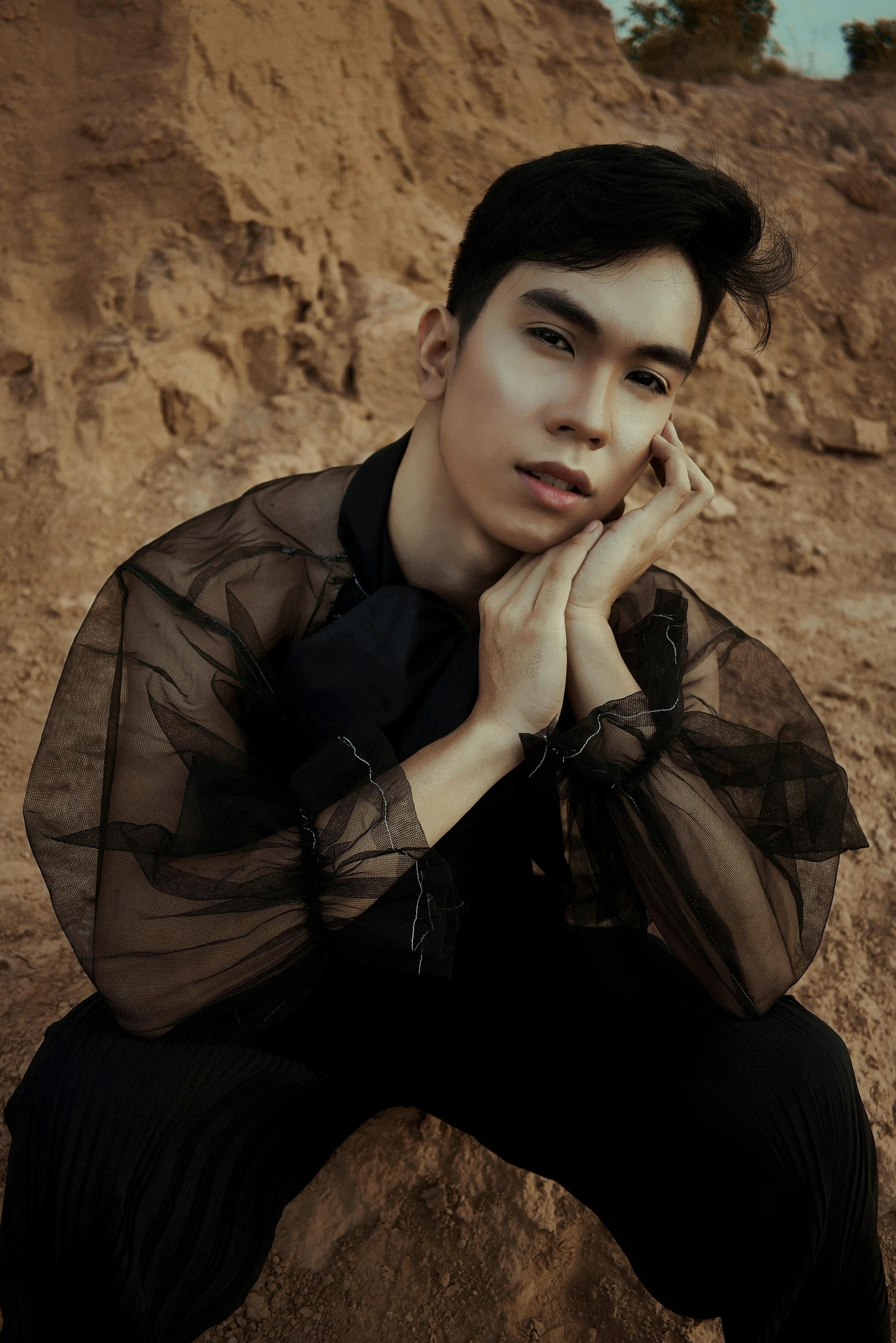 Portrait of a young man in a sheer lace top sitting outdoors with a thoughtful expression.