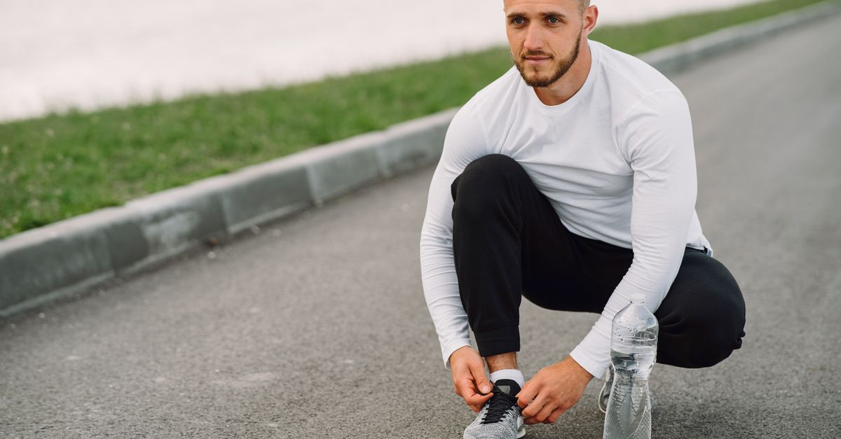 A man in athletic wear ties his shoe on a road, ready for exercise.