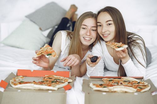 Free Two young women enjoying pizza and watching TV together on a cozy bed. Stock Photo