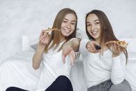 两名年轻女子在舒适的室内环境中，微笑着享用披萨，并指着遥控器
