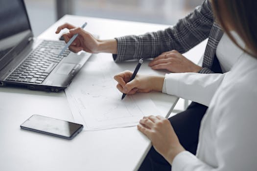 Two professionals collaborating on business plans using a laptop and paper documents.