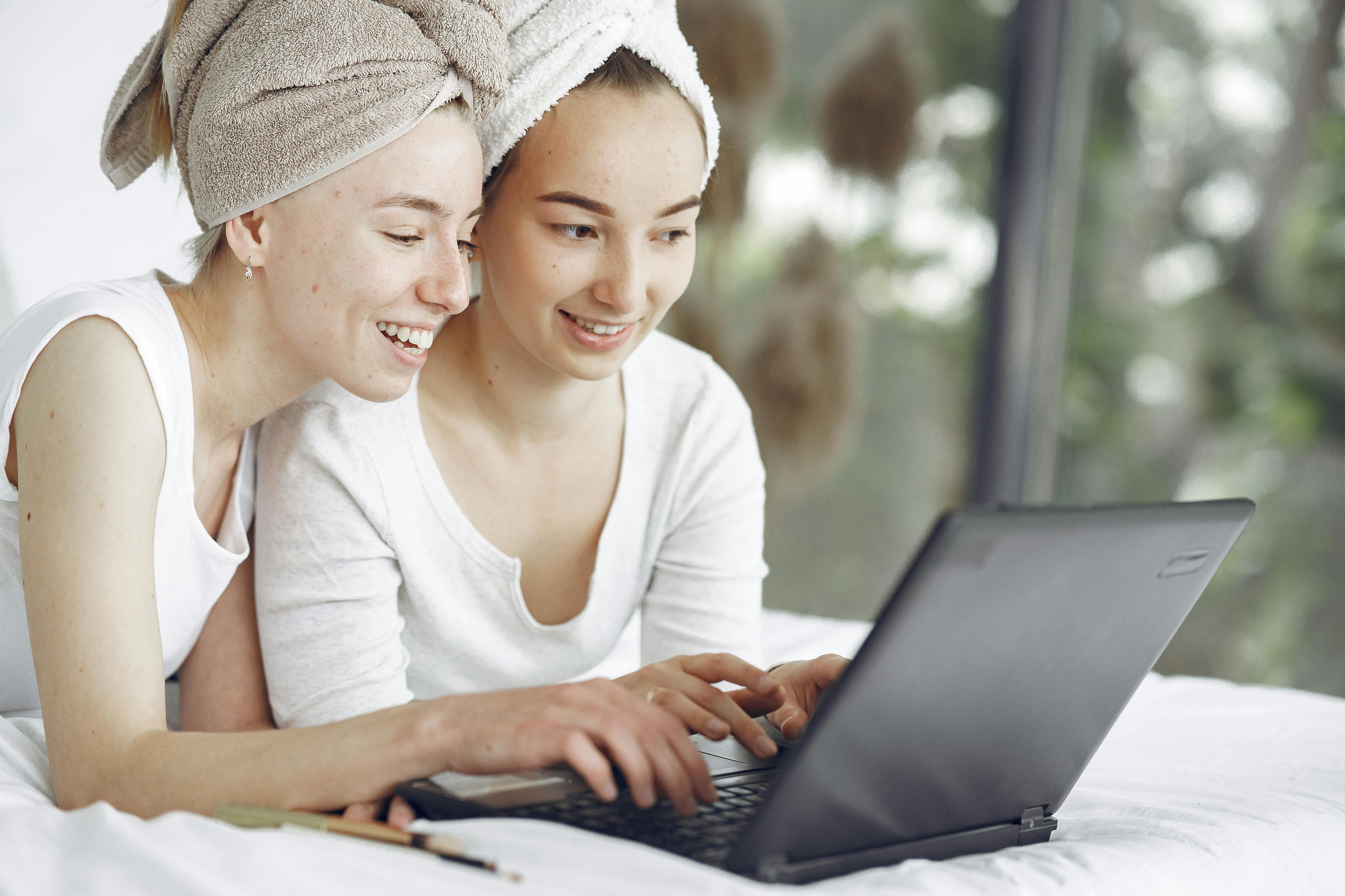 Two young women enjoying leisure time together, using a laptop indoors with towels on their heads.