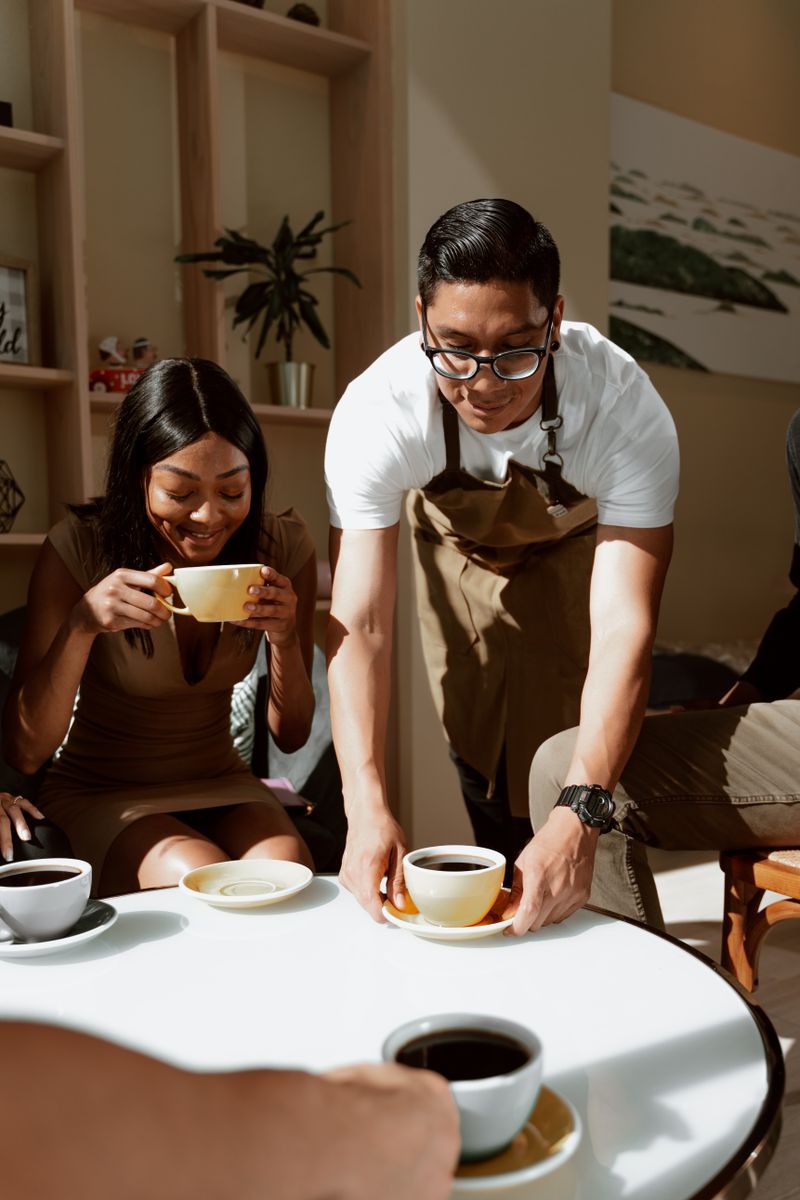 A barista setting a cup of filter coffee on a cafe table while a guest sips hers — photo by RDNE Stock project on Pexels