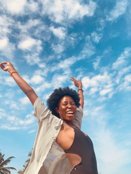 Side view of expressive young African American female tourist with curly hay in stylish swimsuit enjoying summer day with raised arms on beach and smiling
