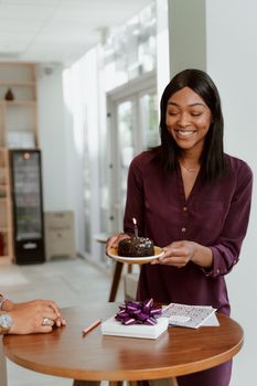 Woman holding a chocolate cake with a candle, celebrating indoors with a gift on the table.