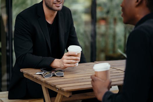 Two adults enjoying a coffee meeting outdoors, holding disposable cups on a wooden table.