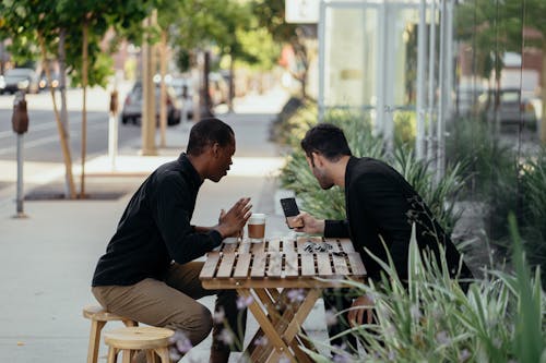 Two Men Sitting by the Wooden Table While Using a Smartphone