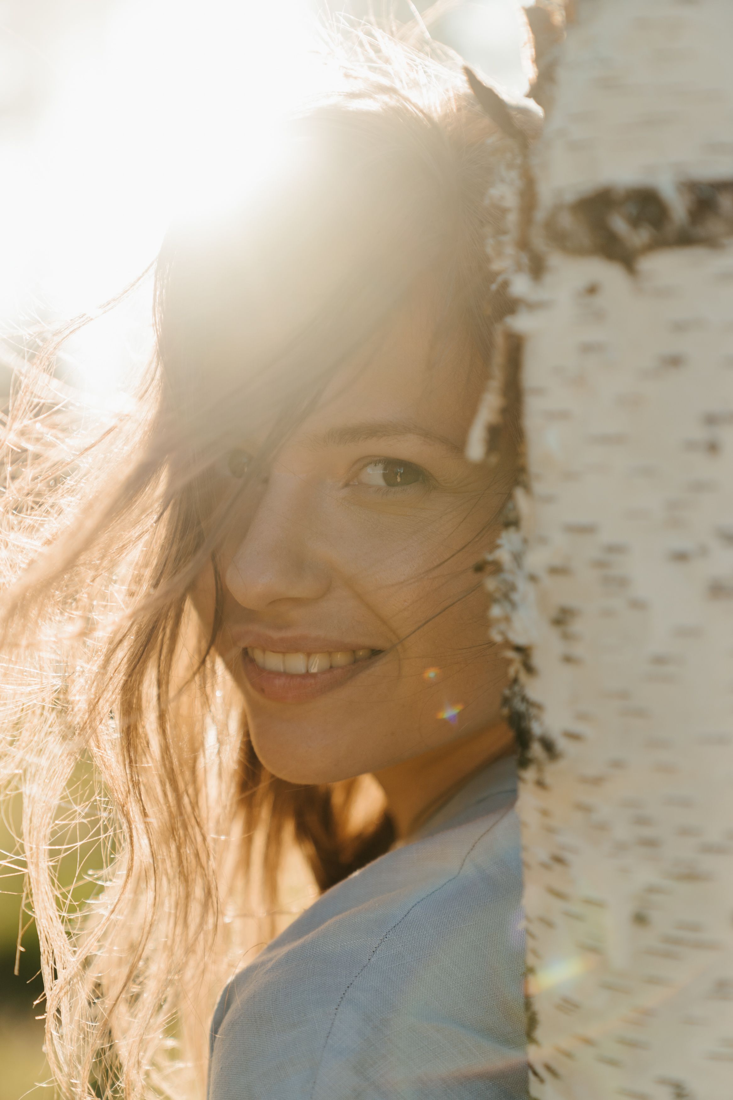 A woman smiles in the sunlight beside a birch tree, capturing a joyful outdoor moment.