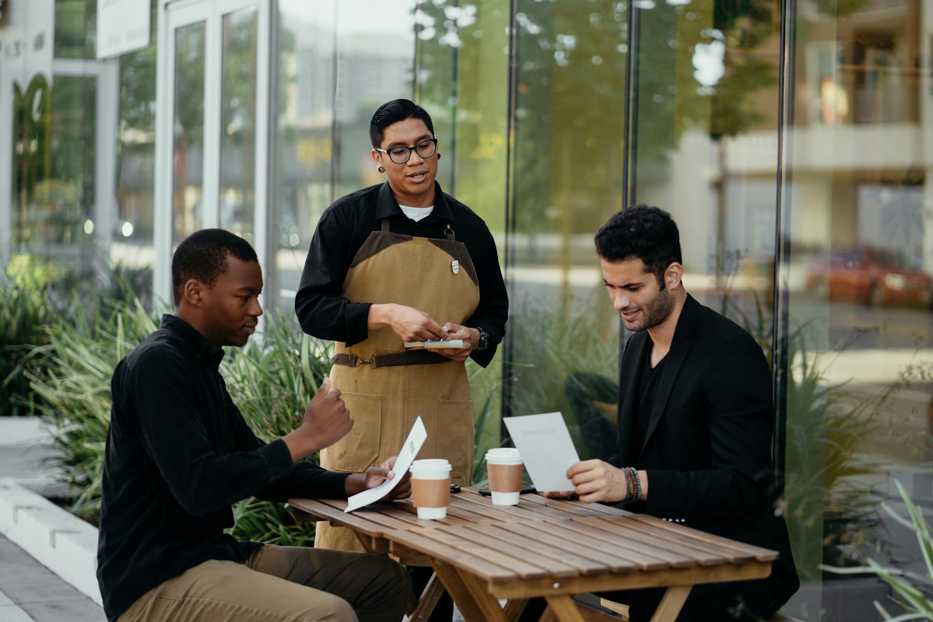 Two customers engaging with a waiter at an outdoor café table, reviewing the menu.