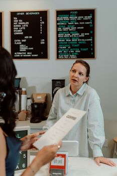 A woman at a café counter engaging with a customer holding a menu. Beverage and food menu visible.