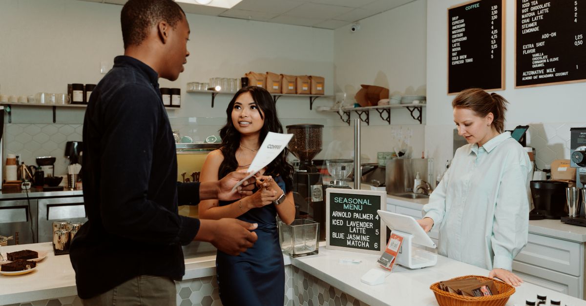 Dois clientes interagem com um barista enquanto fazem pedidos em um balcão de café moderno.