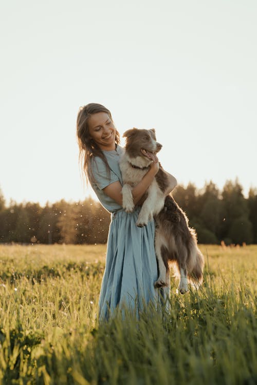 Free Woman in Blue Dress Standing on Green Grass Field With White and Black Long Coated Dog Stock Photo