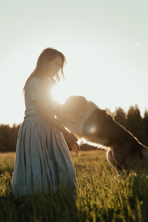 Free Woman in White Long Sleeve Dress Standing Beside Brown Dog on Green Grass Field Stock Photo