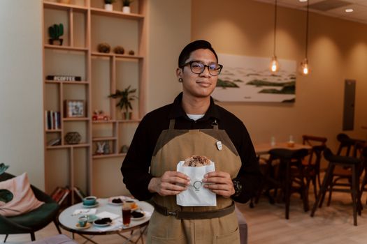 A barista in an apron holds chocolate chip cookies inside a cozy coffee shop.