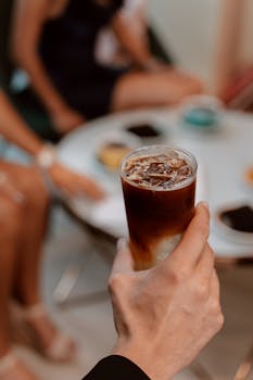 A hand holds an iced coffee during an indoor cafe gathering, featuring a blend of lively conversation and modern style.