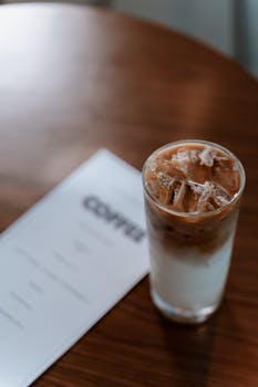 Tall iced coffee with milk, placed on a wooden table next to a blurred menu.