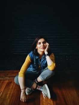 Young woman in casual attire sitting indoors against a dark brick wall.