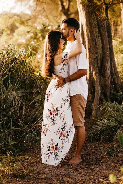 Free Side view full body serene boyfriend embracing and kissing stylish girlfriends forehead while standing together in abundant summer forest Stock Photo