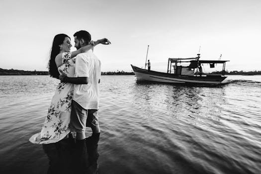Black and white full body smiling young couple in stylish summer clothes embracing while standing in seawater near boat and looking at each other with tenderness