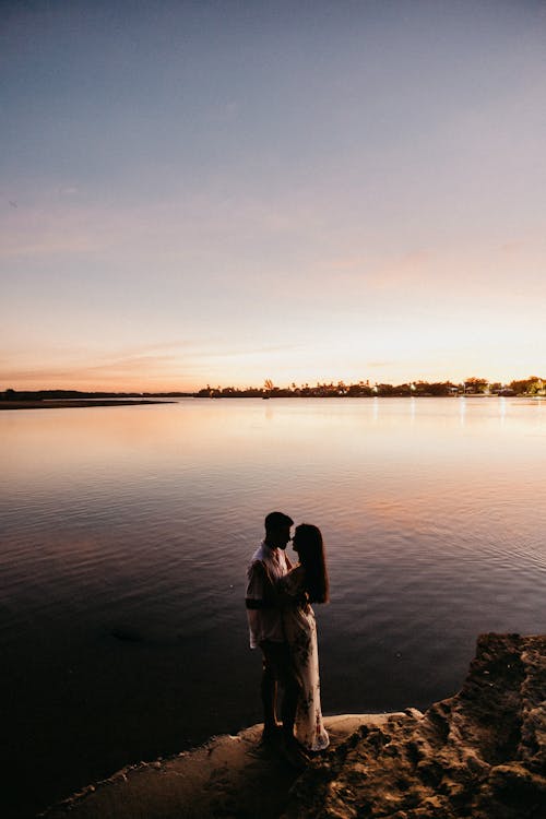 Side view unrecognizable couple bonding gently while standing together on scenic river shore in tranquil sunset time