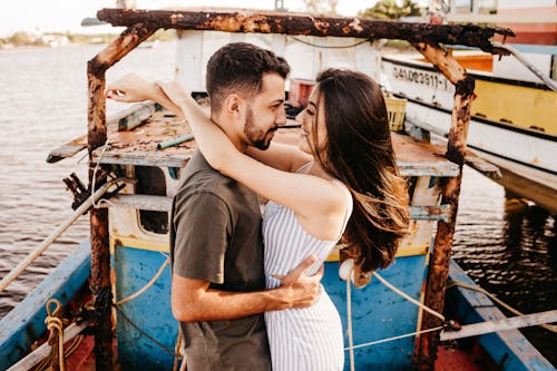 Side view cheerful young couple in stylish summer wear bonding and looking at each other with love while standing on old boat floating on river