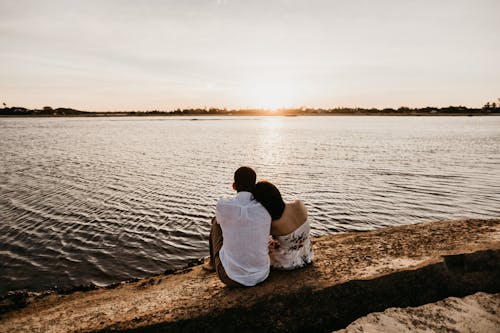 Back view unrecognizable female resting head on boyfriends shoulder while sitting together on stony river coast during romantic sunset