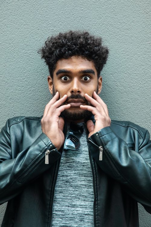 Free Young man in a black leather jacket looking surprised with hands on face, against a neutral background. Stock Photo