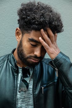 A young man with curly hair in a leather jacket appears thoughtful outdoors.