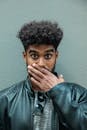一名卷发遮嘴的男子穿着皮夹克，面露惊讶，背景中性