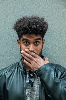 一名卷发遮嘴的男子穿着皮夹克，面露惊讶，背景中性