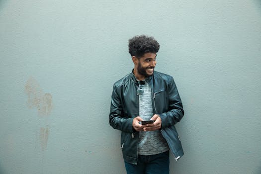 Smiling man in black leather jacket using smartphone against a wall outdoors.