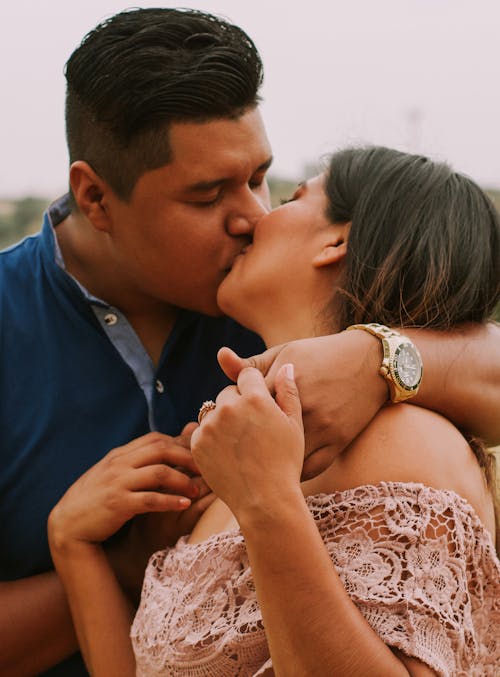 Hispanic man in blue shirt with golden wristwatch kissing girlfriend wearing pink lace dress