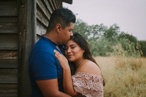 Hispanic man kissing head of smiling wife while standing near gray wooden wall of old building