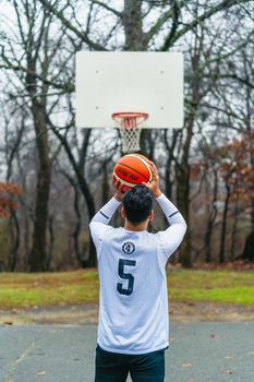 A man shooting a basketball outdoors on a gloomy day, aiming at a hoop.
