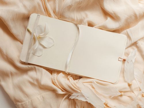 Free An open sketchpad adorned with a white flower, laid on a silky fabric with delicate feathers. Stock Photo