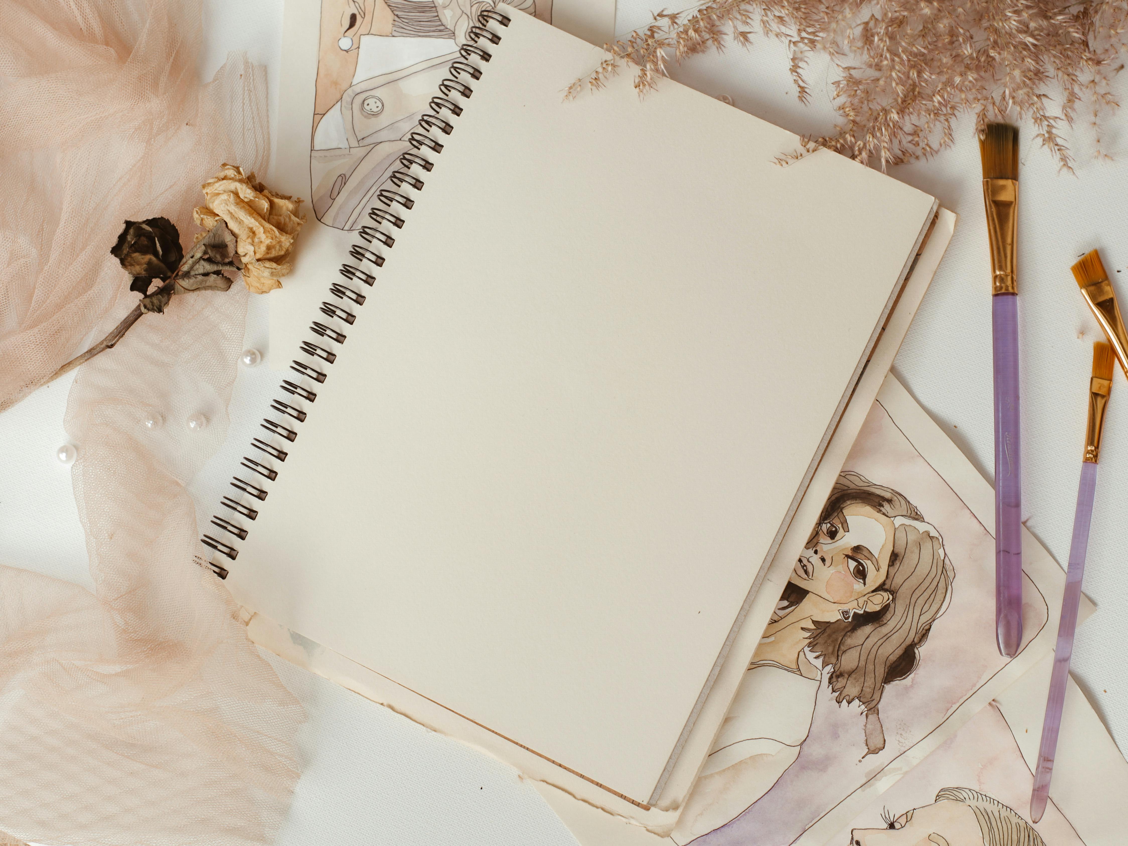 Elegant composition of art supplies including a sketchbook, paintbrushes, and dried flowers on a white surface.