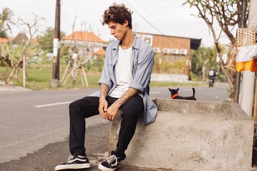 Thoughtful young man with tattoos sits outdoors in casual clothing, exuding a relaxed vibe.