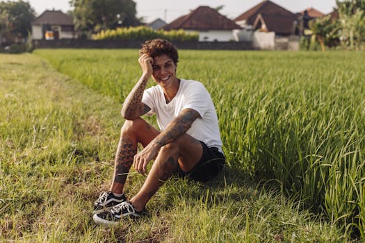 Smiling young man with tattoos sitting in a lush green rice field under the sun.