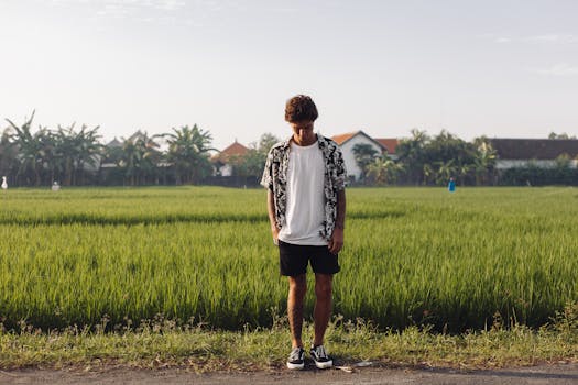 Young man in casual attire standing in a vibrant green field with distant houses.