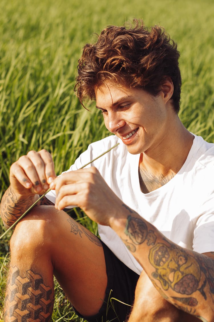 Young Man With Tattoos Sitting On A Field With A Piece Of Grass In His Mouth And Smiling 