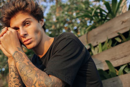 Portrait of a young tattooed man with brown hair sitting outdoors, showcasing inked arms and a nose ring.
