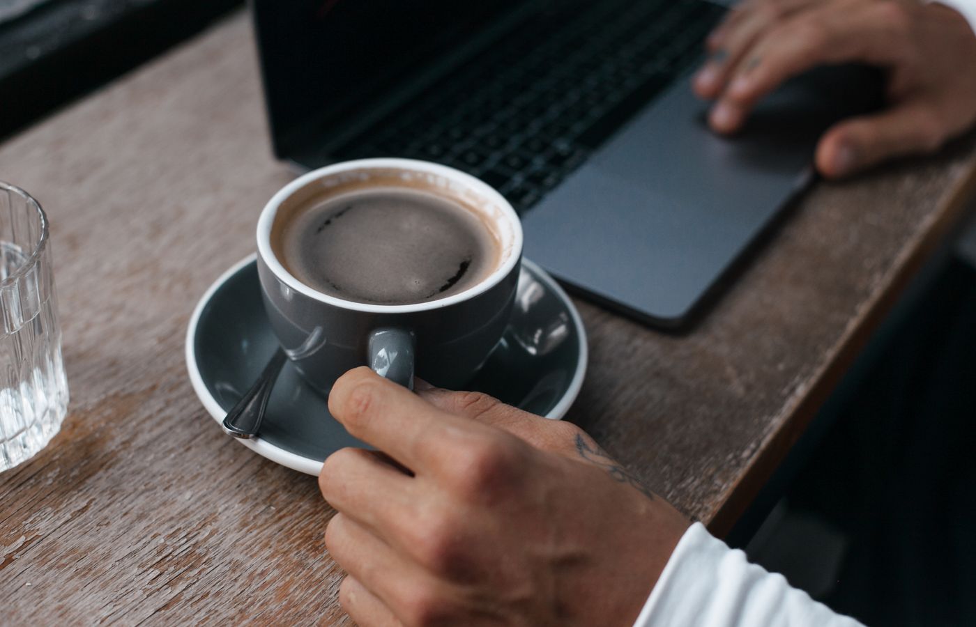 A person holding a coffee cup while working on a laptop at a wooden table.