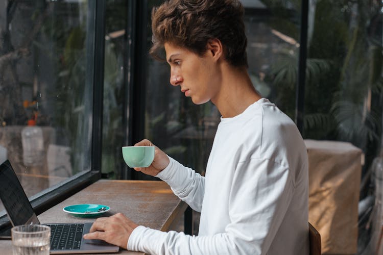 Young Man Working On A Laptop, Drinking Coffee In A Cafe 