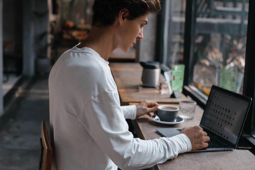 A young man sitting in a café working on a laptop beside a cup of coffee — photo by ROMAN ODINTSOV on Pexels