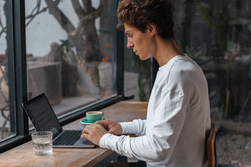 Free A young adult in white long sleeves works on a laptop by a cafe window, enjoying a serene atmosphere. Stock Photo