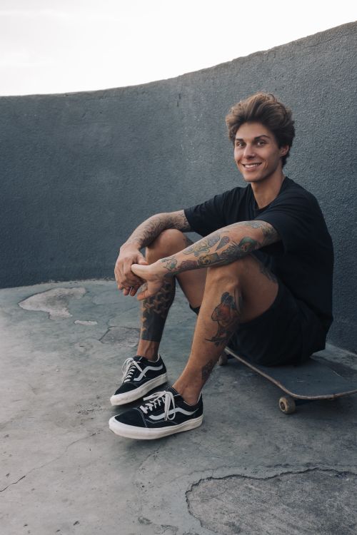 Free Smiling young man with tattoos sitting on skateboard in urban setting. Stock Photo