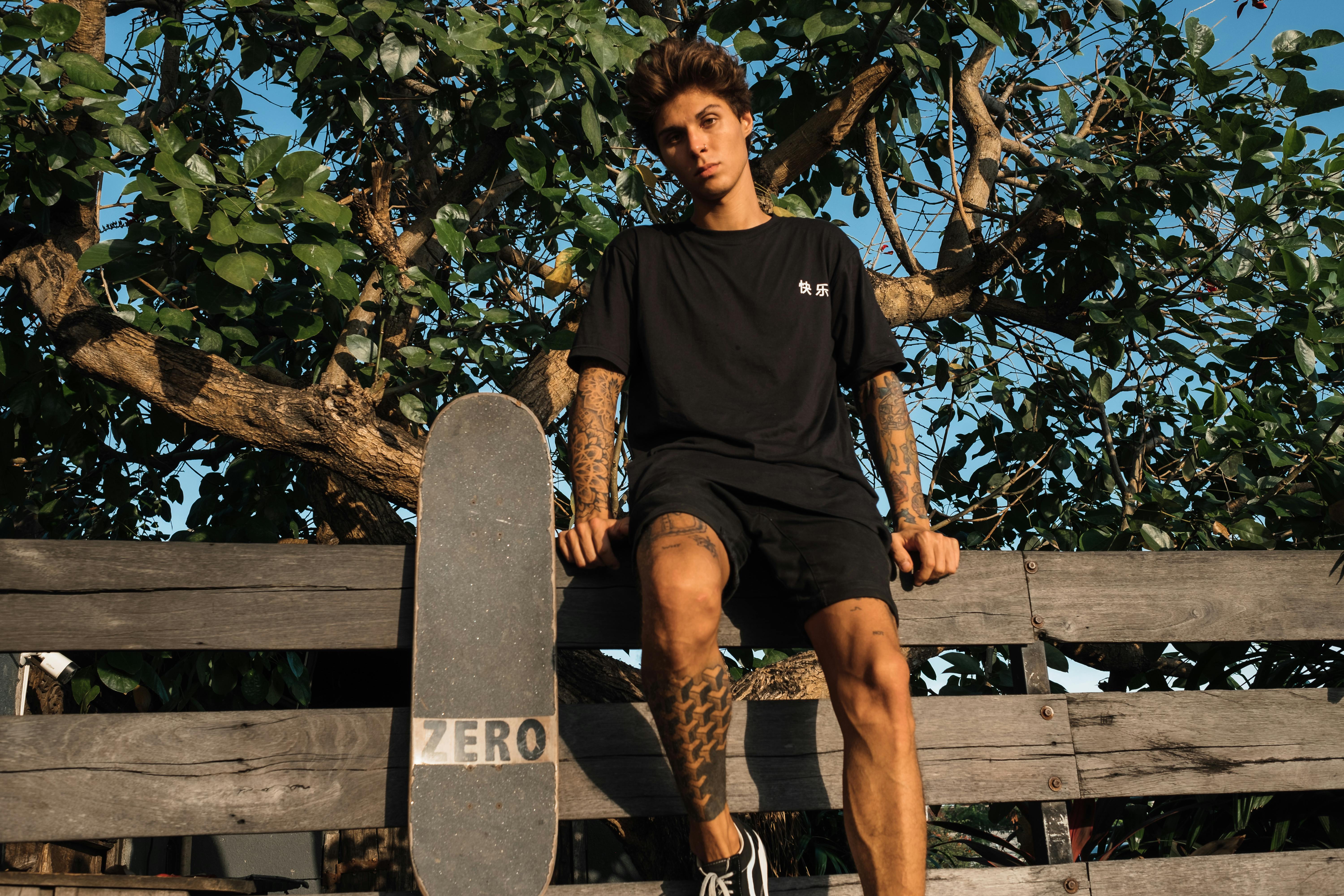 一位穿着黑色服装、身上有纹身的年轻滑板手，随意地坐在树下质朴的木栅栏上