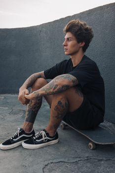 一位穿着黑色服装、纹身的年轻滑板手若有所思地坐在户外的滑板上