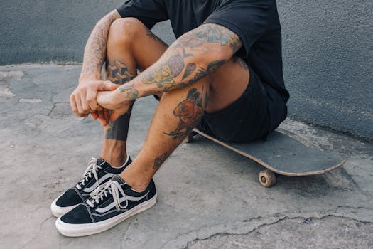 A tattooed man sits casually on a skateboard against an urban concrete backdrop.