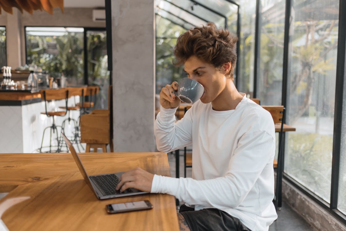 Man in White Long Sleeve Shirt Holding Black Ceramic Mug Sitting Beside Brown Wooden Table